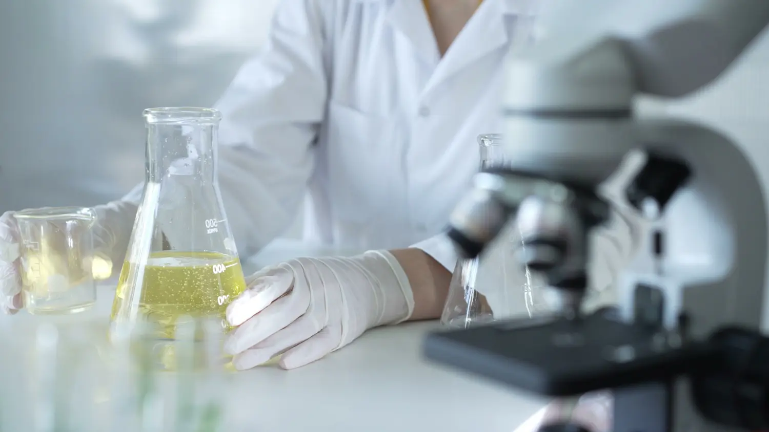 Ein Wissenschaftler, der einen Laborkittel und weiße Schutzhandschuhe trägt, gießt eine gelbe ölige Flüssigkeit von einem Becherglas in ein anderes in der Nähe eines Mikroskops im Labor, Nahaufnahme. Medizin- und Wissenschaftskonzept